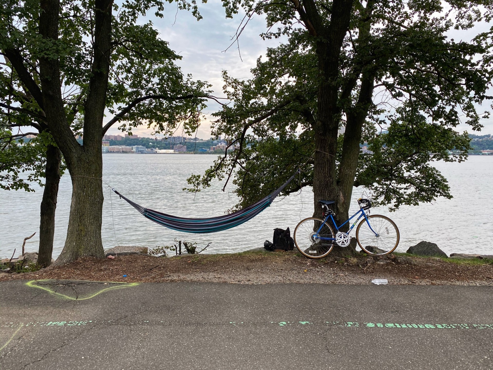 A hammock swings between two trees with a river in the background. A bike is parked at one tree. 