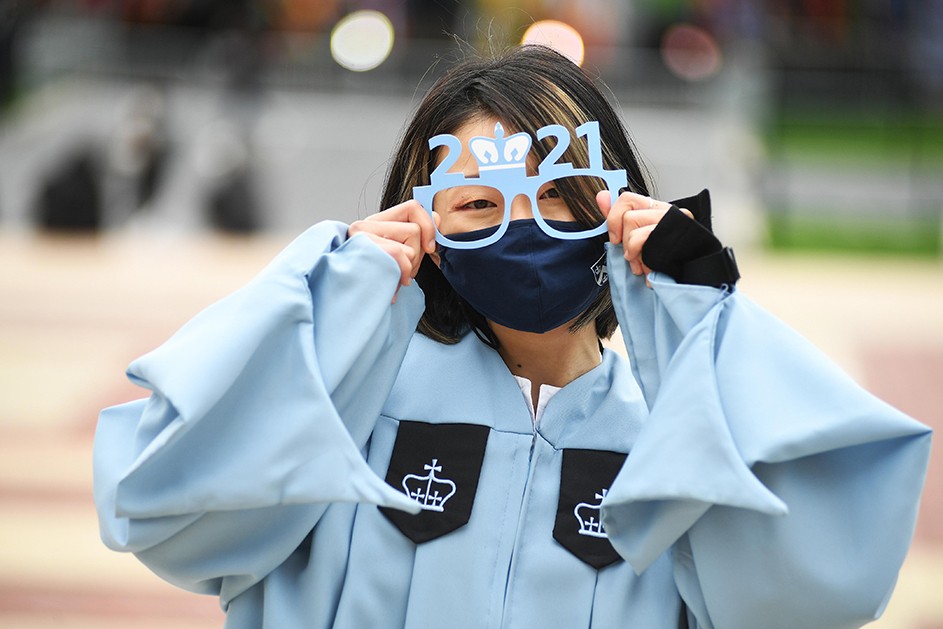 A woman in a blue robe holds up paper 2021 graduation glasses. 