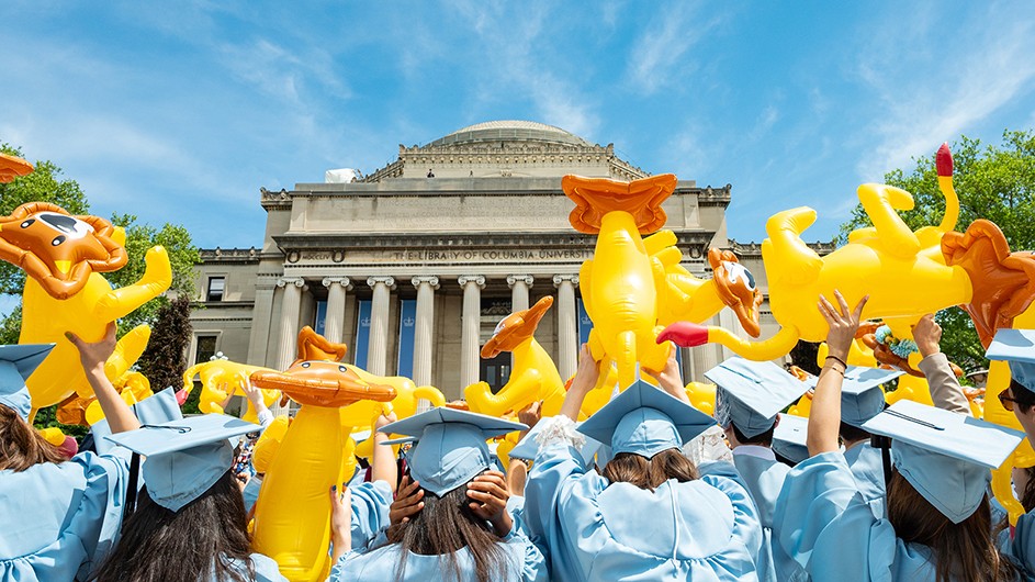 Students in graduation gowns wave plastic lions at Low Library.