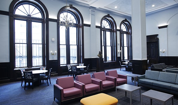 The renovated Walter and Shirley Fan (BUS'93) Wang Lounge at Columbia University with couches, tables, and chairs, against the backdrop of big brightly-= lit windows. 
