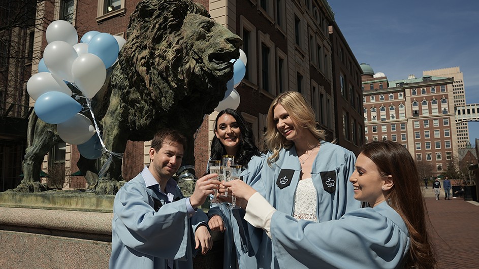 Four graduates toast in front of Scholars Lion