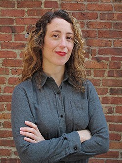 A woman wearing a gray, button-down shirt with long, curly hair and bright lipstick stands with folded arms before a brick wall.