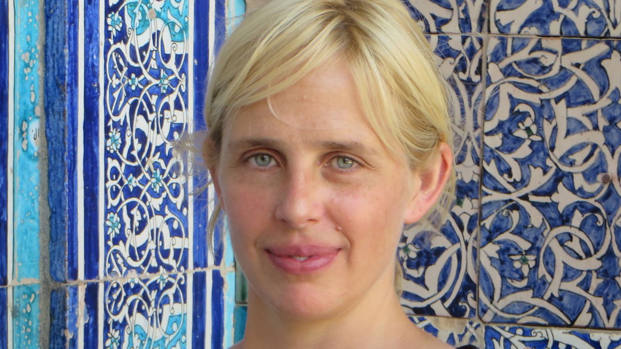 A woman with blond hair against a background of blue and turquoise Islamic tilework.