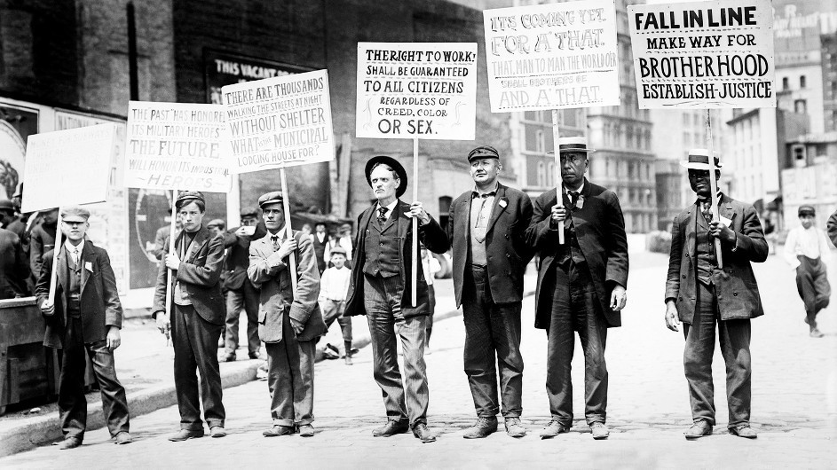 A black and white vintage photo from 1909 of a group of men dressed in jackets and ties walking on a New York City street men holding up protest signs about unemployment and the need for jobs.