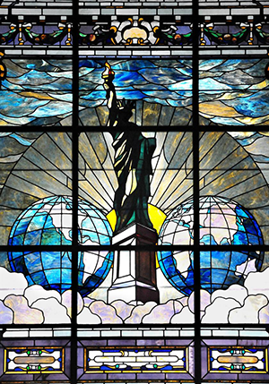 Stained glass window depicting two worlds and the Statue of Liberty on clouds in front of a rising sun.