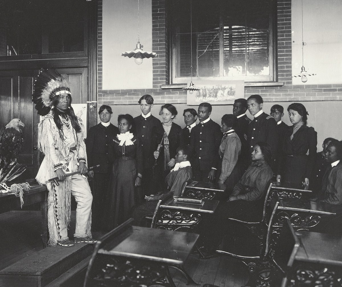 A Native American man wearing a feathered headdress stands in a classroom of students.