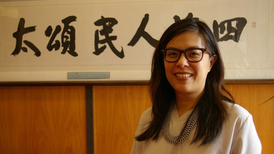 Eugenia Lean: A bespectacled Asian woman with long dark hair and wearing a white shirt and a beaded necklace, smiles in front of a framed piece of Chinese calligraphy