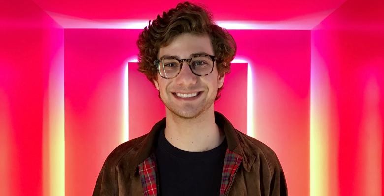 image of jonny cohen dressed in a dark top wearing dark eyeglasses in front of a neon pink and yellow backdrop