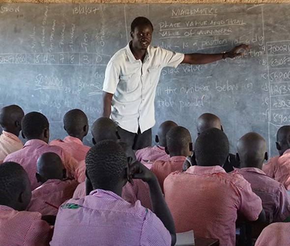 A teacher in white shirt at a chalkboard with writing on it stands in front of students with with red shirts and their backs to the camera.