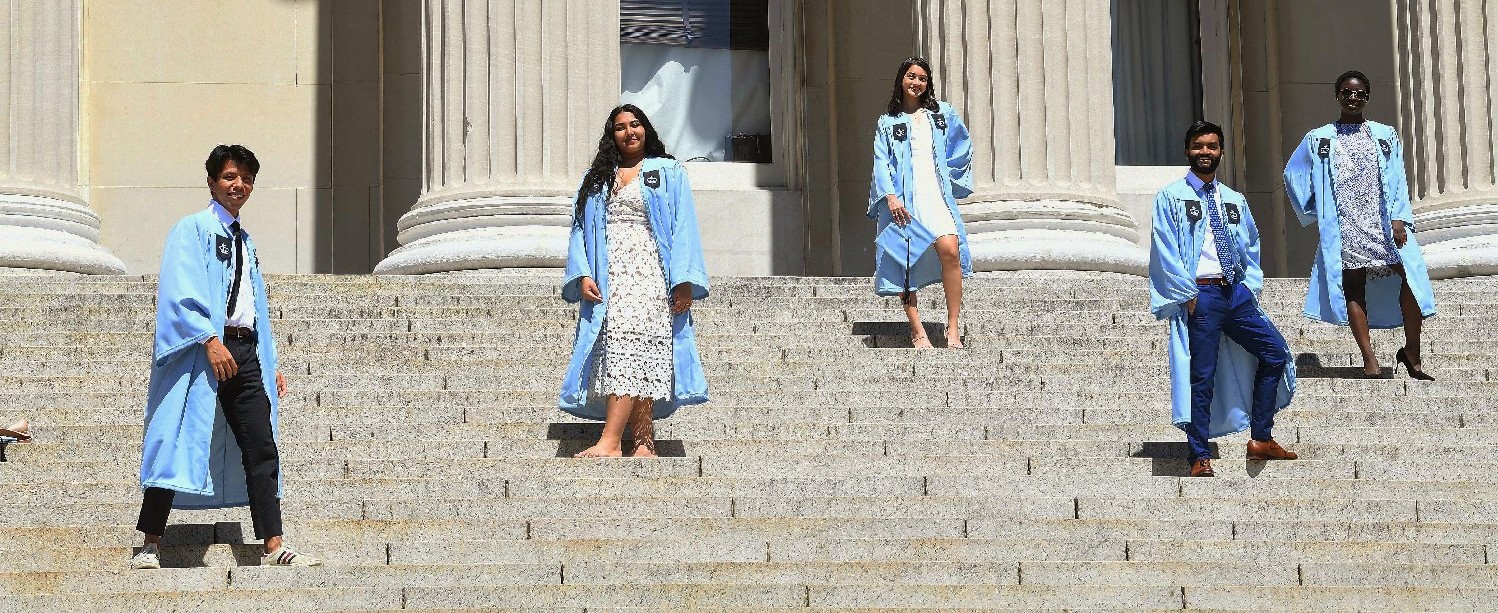 A diverse group of graduates in their Columbia gowns stand at a social distance on the steps of Low Library