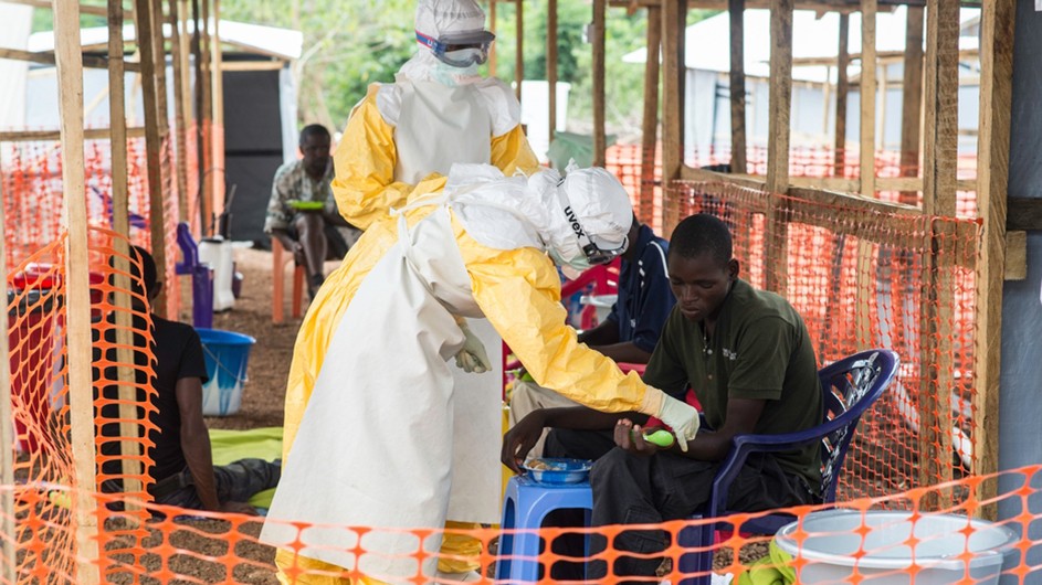 Two medics in yellow and white uniforms with protective eye and head gear treat an African young man with Ebola