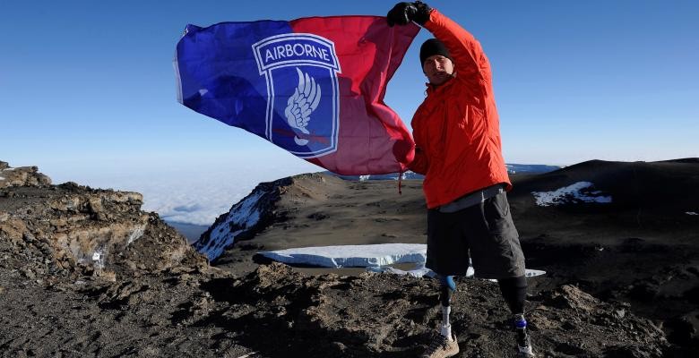 neil duncan waving a red and blue flag while standing atop a mountain dressed in red jacket and dark bottoms