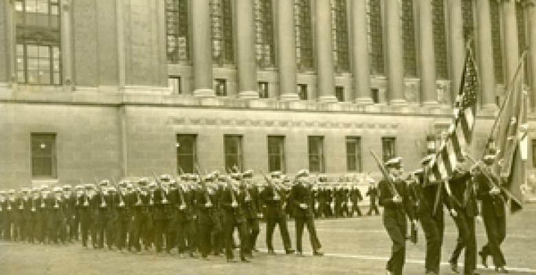 Men dressed in naval uniforms march across the grounds in front of Butler Library.