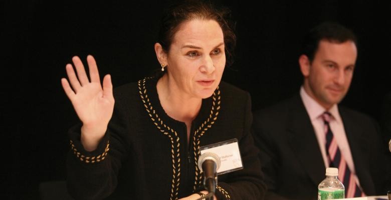 Speaking into a microphone while seated at a table, a woman raises her right hand, gesturing