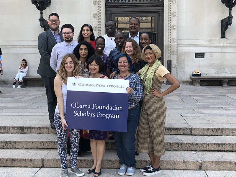 A group of 14 women and men stand on the steps in front of Butler Library. The front row of three women holds a placard reading "Columbia World Projects, Obama Foundation Scholars Program."
