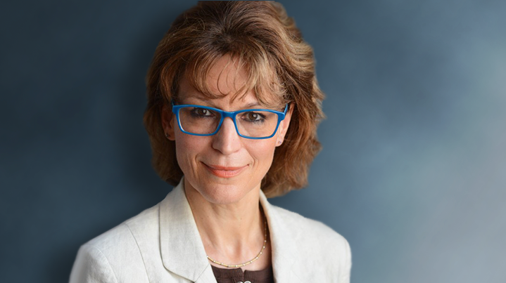 Agnes Callamard in blue glasses, a white jacket in front of a blue background. 
