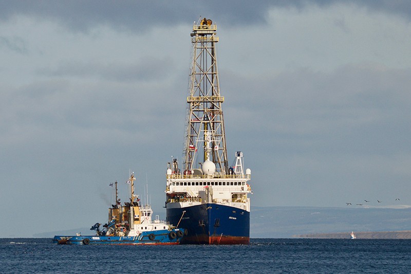 A research vessel with a tower on it is pulled by a tugboat on the ocean. 
