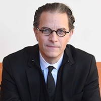 Man with round-framed glasses in dark suit & tie.