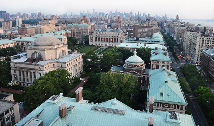 An aerial view of the Morningside Campus. The dome-shaped roof of Low Library risinig above full trees and the New York City skyline faded in the backdrop of the photo.