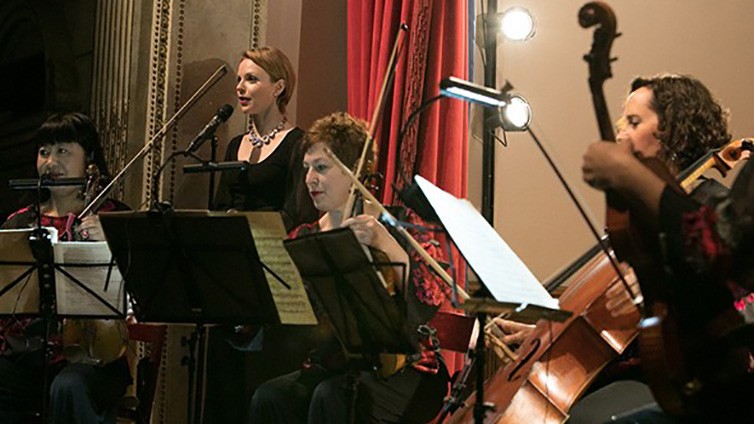 The Cassat String Quartet sit with their instruments in front of their music stands on stage with a red curtain to their left and Magdalena Baczewska at a microphone.