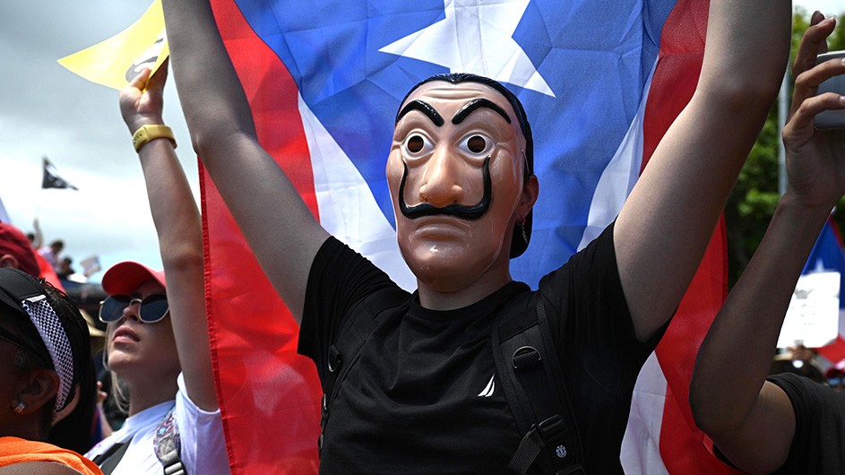 A protestor holds up a Puerto Rican flag and wears an Anonymous-like mask. 