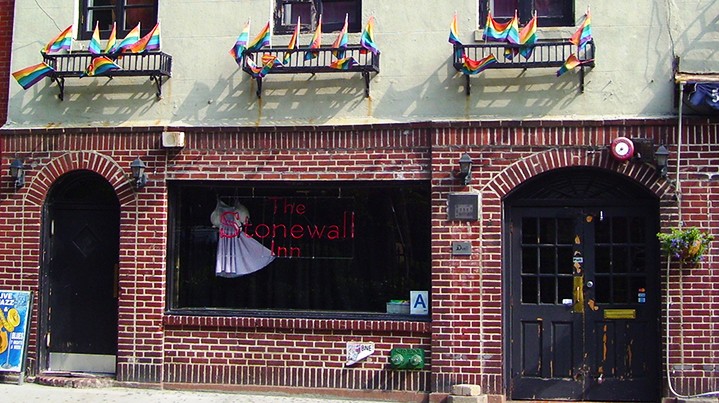 The red brick storefront of The Stonewall Inn with three windows above lined with rainbow flags. 