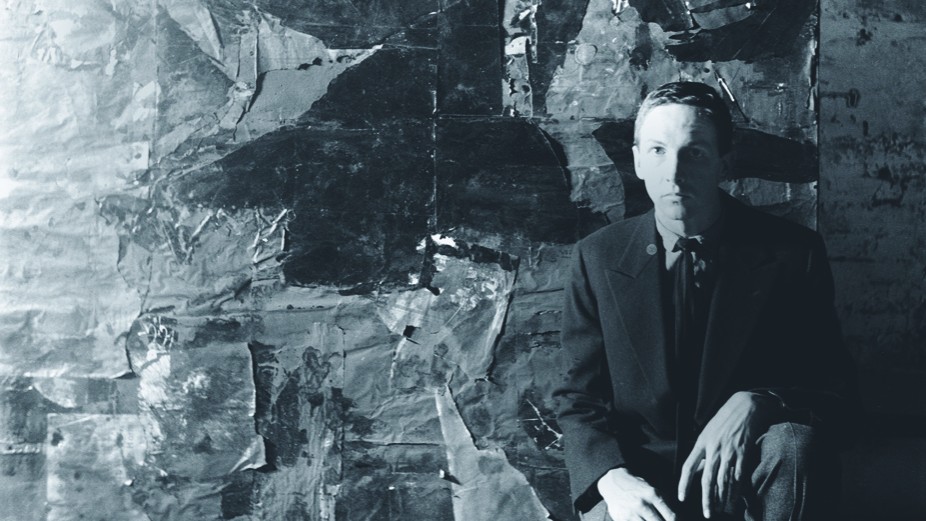 The artist Robert Rauschenberg, a man with close-cropped hair, is seated in front of one of his artworks