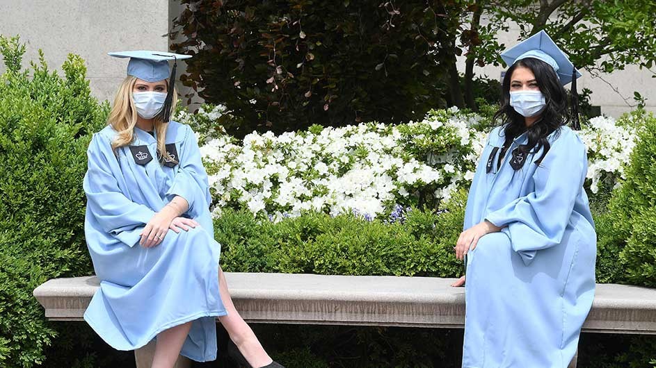 Two women in Columbia blue caps and gowns and face masks sit on a bench on campus.