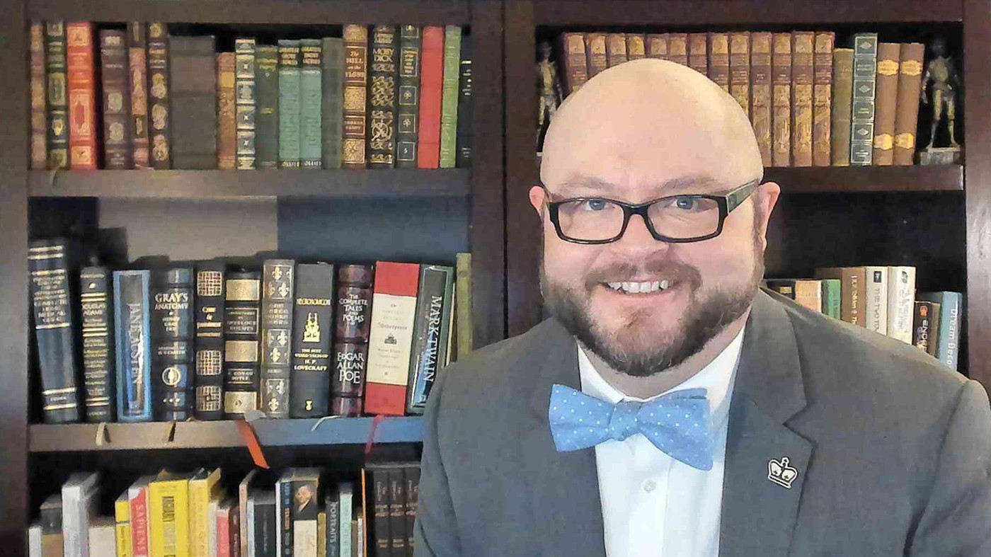 Allen Darrah: A bald man with a mustache and beard and dark-framed glasses wearing a blue bowtie, white collared shirt and jacket with a lapel pin shaped like a crown, stands before book-lined shelves.