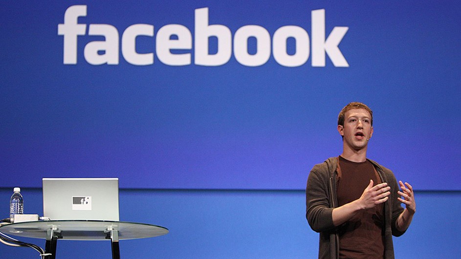 Mark Zuckerberg, in a brown t-shirt and hoodie, stands in front of a large blue backdrop with Facebook printed across the top. There is a high, round, glass-top table holding a laptop and water bottle across the stage from him.