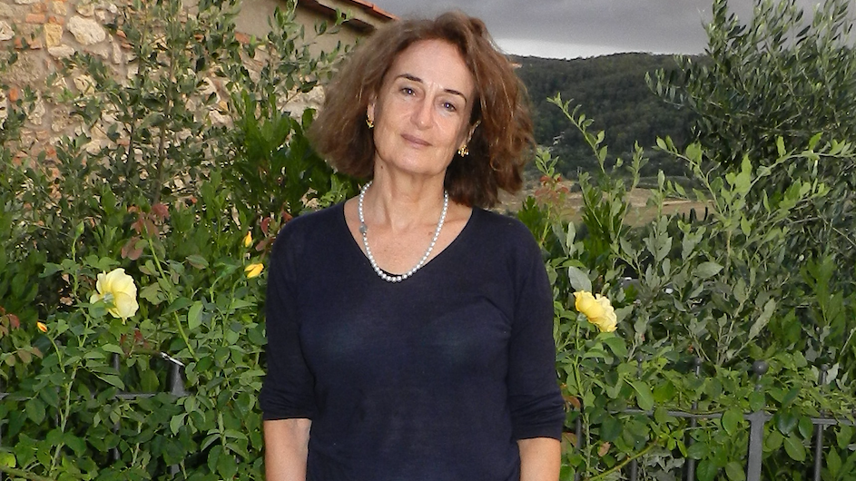 An image of a woman with shoulder length brown hair wearing a dark blue v-neck sweater standing in a garden