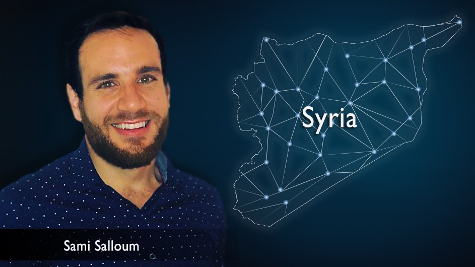 Man with short, dark hair and a dark mustache and beard, in a blue shirt, against the backdrop of a dark blue astral map of Syria. 