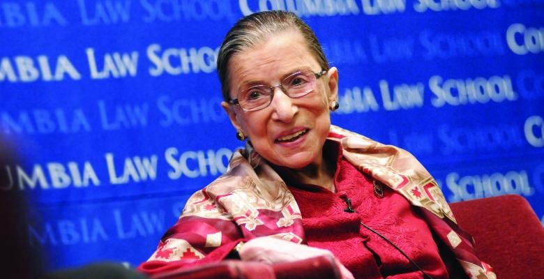 Ruth Bader Ginsburg in a red blouse in front of a blue Columbia Law School poster