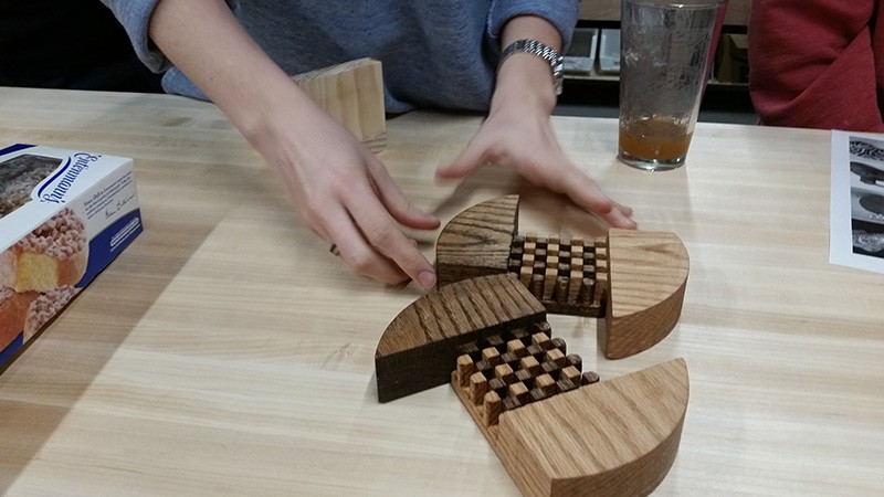 A pair of hands gesticulate toward two wooden boards with elaborate joinery on a table