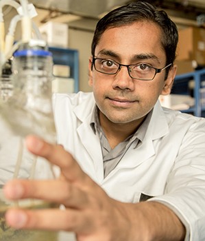 half photo of dark haired man with glasses and white lab coat holding large test vial