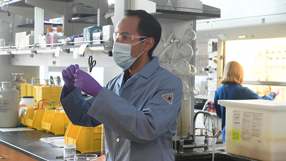 Dark-haired man in mask and safety goggles wearing purple gloves and blue shirt in lab with woman scientist back to him writing on white board