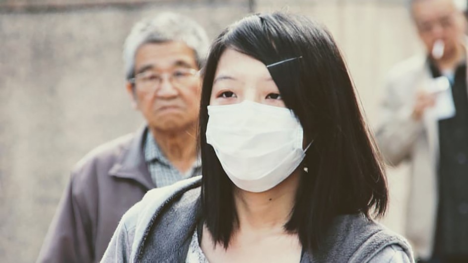 Woman with long black hair wearing white face mask. Older man with grey shirt and grey hair stands behind her 