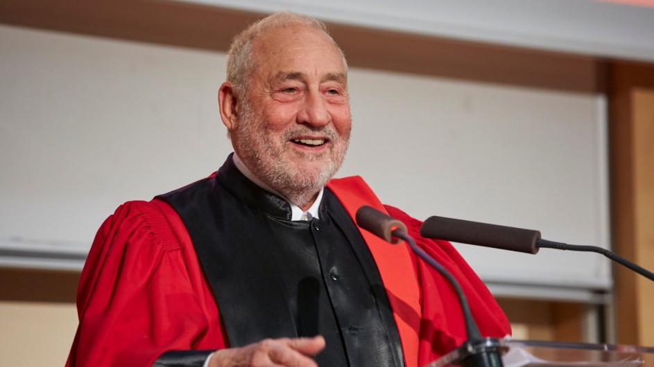 Joseph Stiglitz: a man with gray hair in black and red academic robes, smiling behind a podium