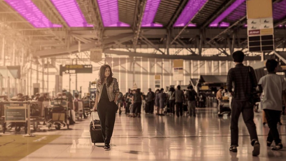 crowded airport terminal, woman with long brown hair, black dress, tan jacket walking under purple lamps, looking like vertical boards on ceiling