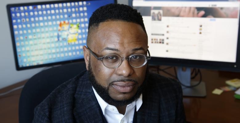 Desmond Patton looking directly into camera dressed in dark blazer white colored shirt.  Dark brown rimmed eyeglasses seated at 2 computer screens located behind him.