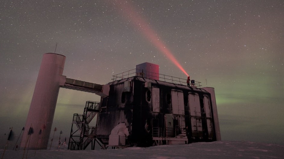 A view of the IceCube Lab at the South Pole with a starry sky above. A winterover is seen on the rooftop shining a headlamp light into the sky. 