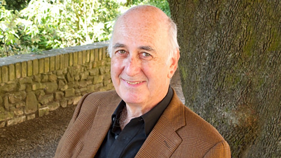 Phillip Lopate: A man wearing a dark shirt and a brown jacket, with white hair, smiles for the camera and stands before trees and a stone wall.