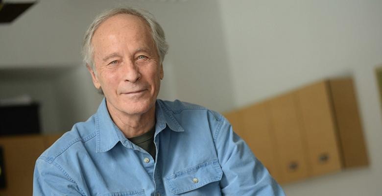 Richard Ford dressed in a blue denim shirt with brown shelves positioned on the wall in the rear