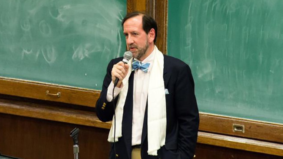 A man wearing a dark jacket and a white scarf holds a microphone and speaks in front of green chalkboards.