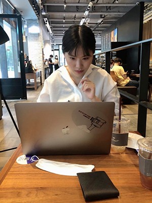 A woman with dark hair and a white shirt works at a laptop on a wooden surface.