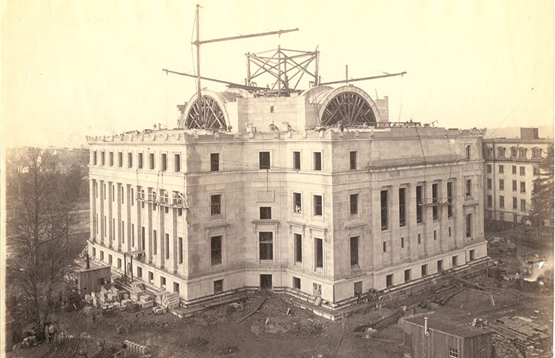sepia toned exterior image of low memorial building while beams are located on the top during construction. men are are at work in the surrounding area of the building