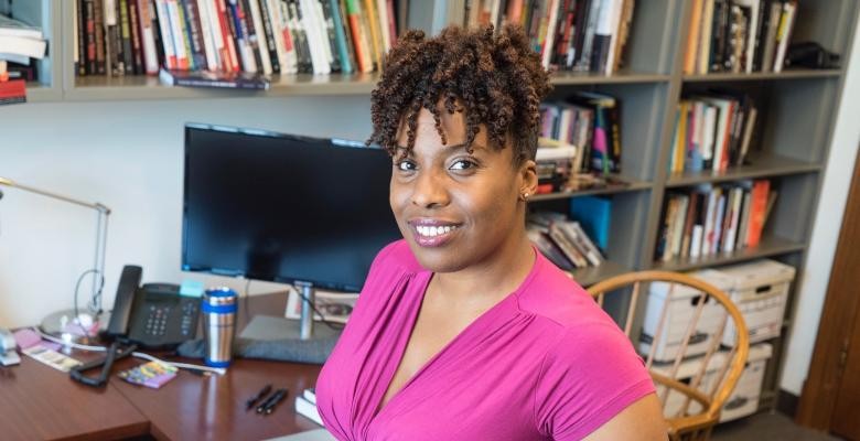 Carla Shedd wears a magenta short sleeved top while seated at a desk with various books placed on shelves behind her.