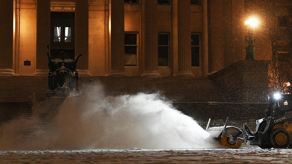 A snow blower blows snow in front of Alma Mater