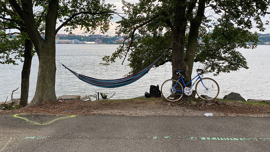 A hammock swings between two trees along New York City's west side of Manhattan, with the Hudson River in the background. A bike is parked at one tree. 