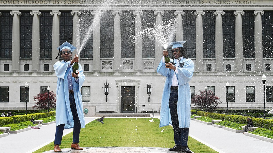Two Columbia students in graduate robes pop champagne in front of Butler Library. 